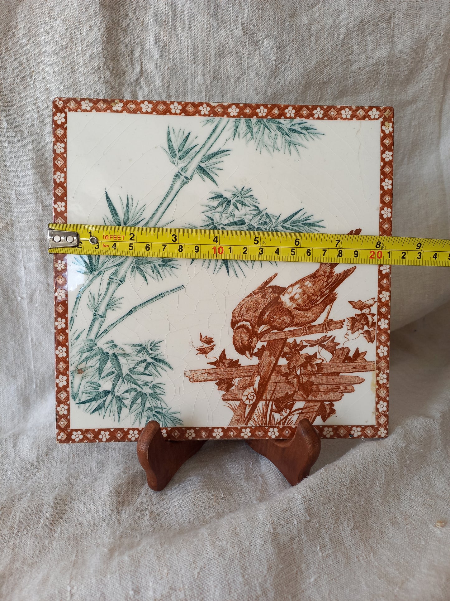 A square ceramic tile with a brown and green  illustration of a bird on a wooden structure, bordered by a floral pattern, shown on a white fabric background with a yellow tape measure for scale.
