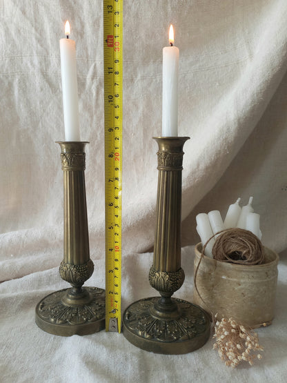 Two ornate brass candlesticks with lit candles on a white fabric background with a tape measure for reference.