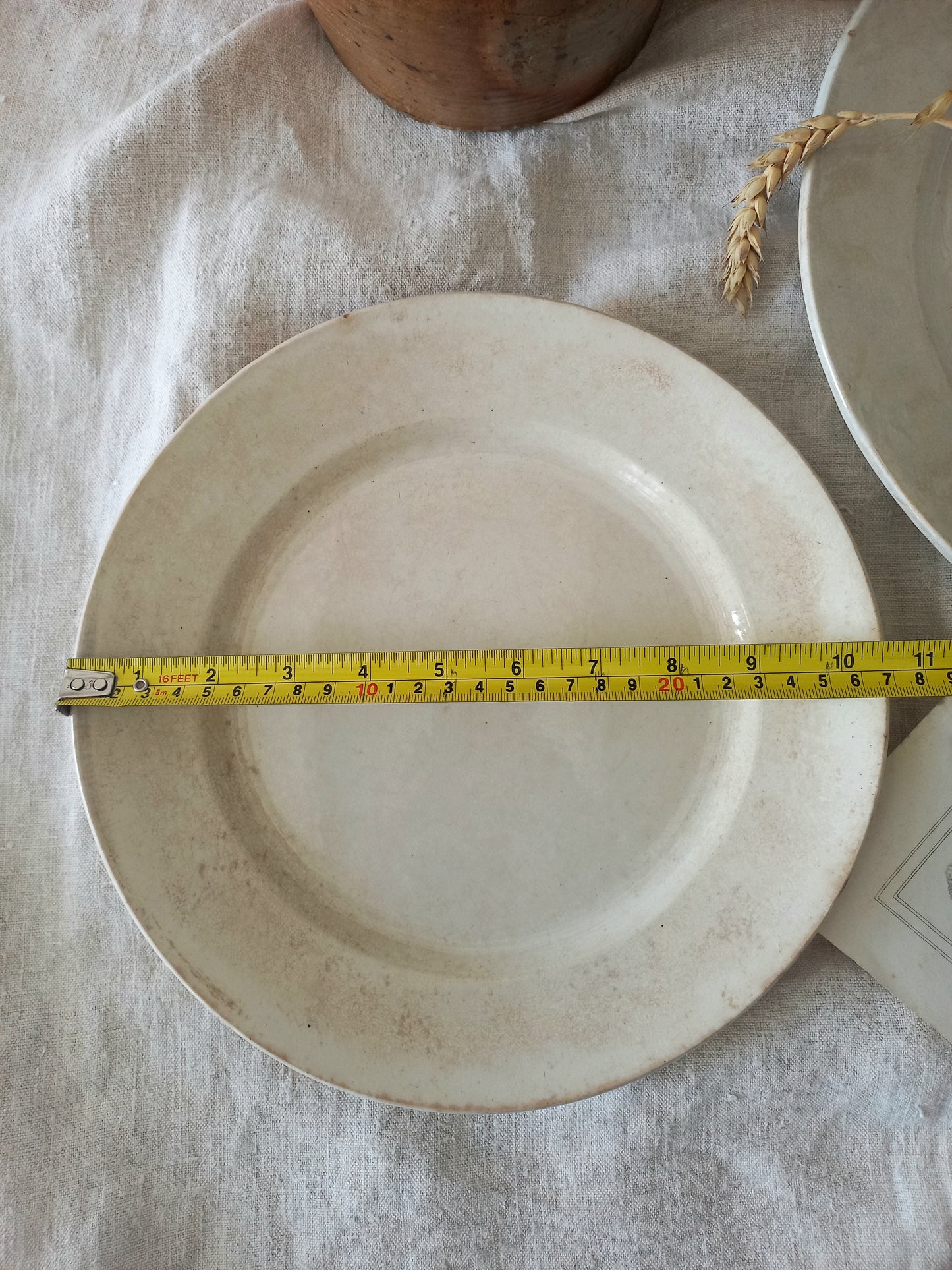 White antique plate with a measuring tape indicating its diameter, on a light fabric background.