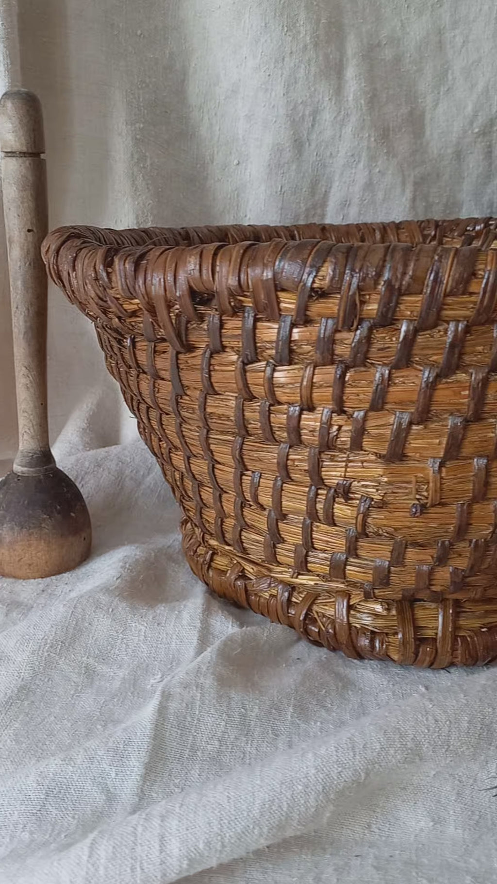 A large vintage round woven vintage basket made of dark brown natural fibers, styled on a draped white linen cloth with rustic wooden kitchen utensils in the background.