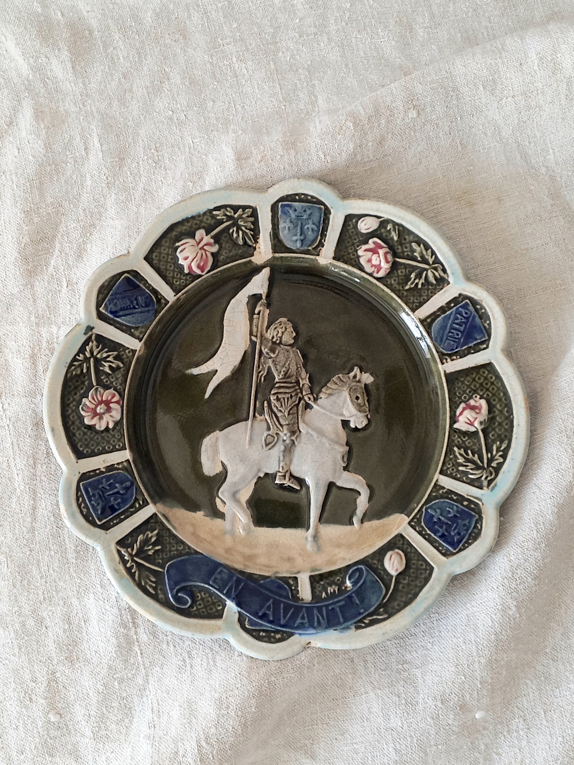 Decorative plate with a scene of Joan of Arc on a horse, surrounded by decorative elements on a white background.