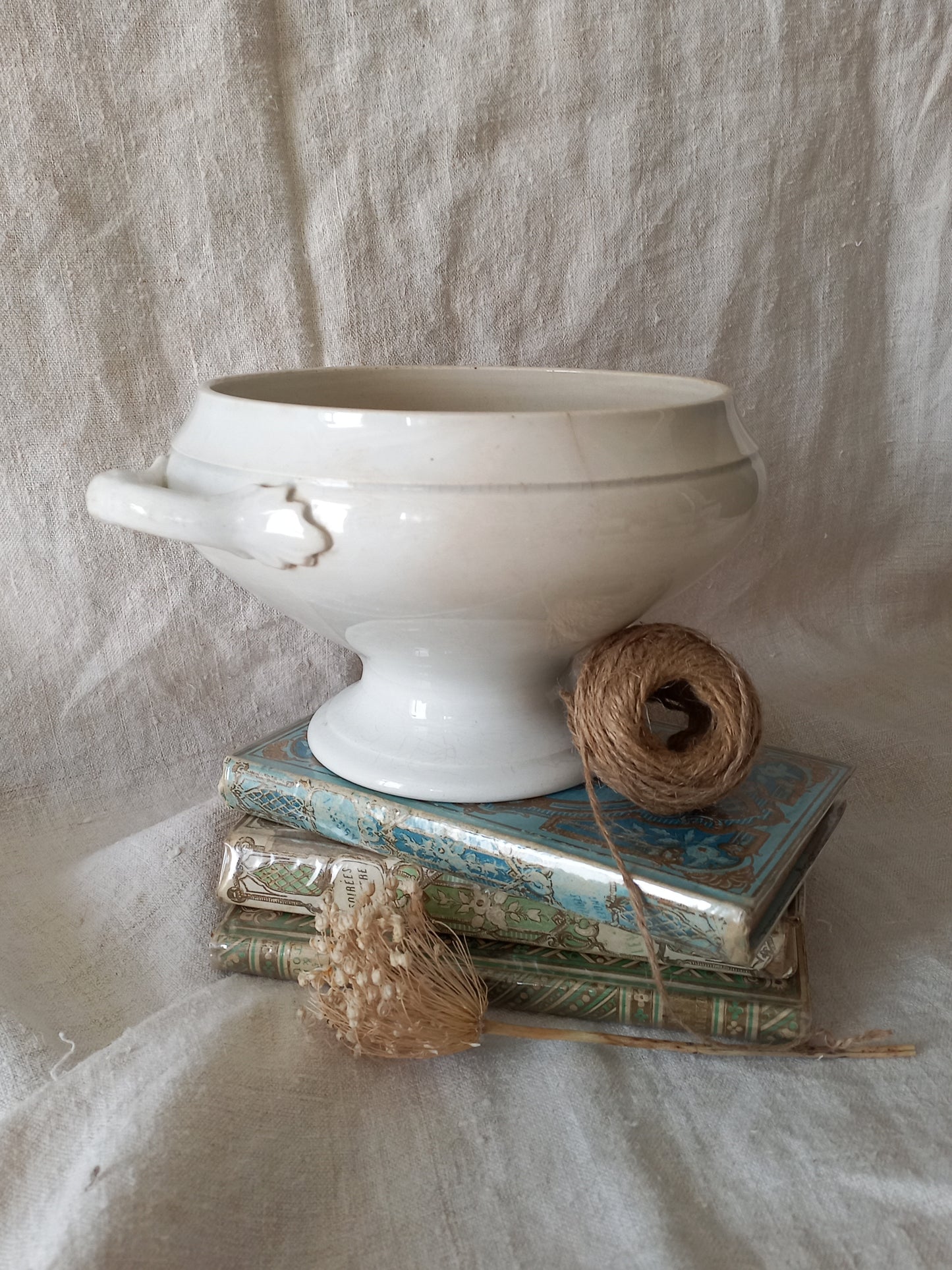 White ironstone bowl on a stack of books with twine and dried flowers against a textured fabric background