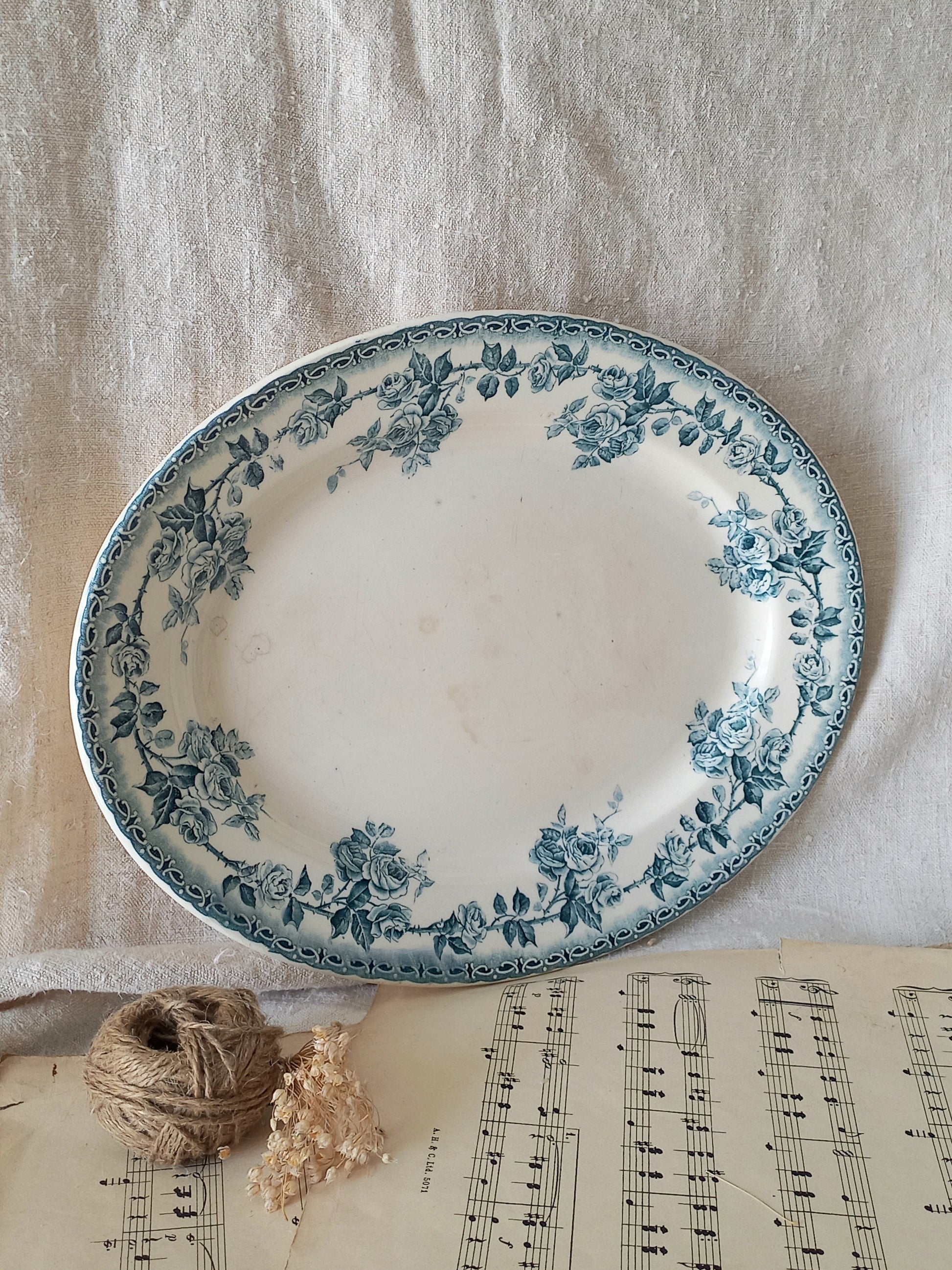 A round white ceramic plate with a blue floral border, styled on a beige linen cloth with a ball of twine, dried flowers, and sheet music underneath.