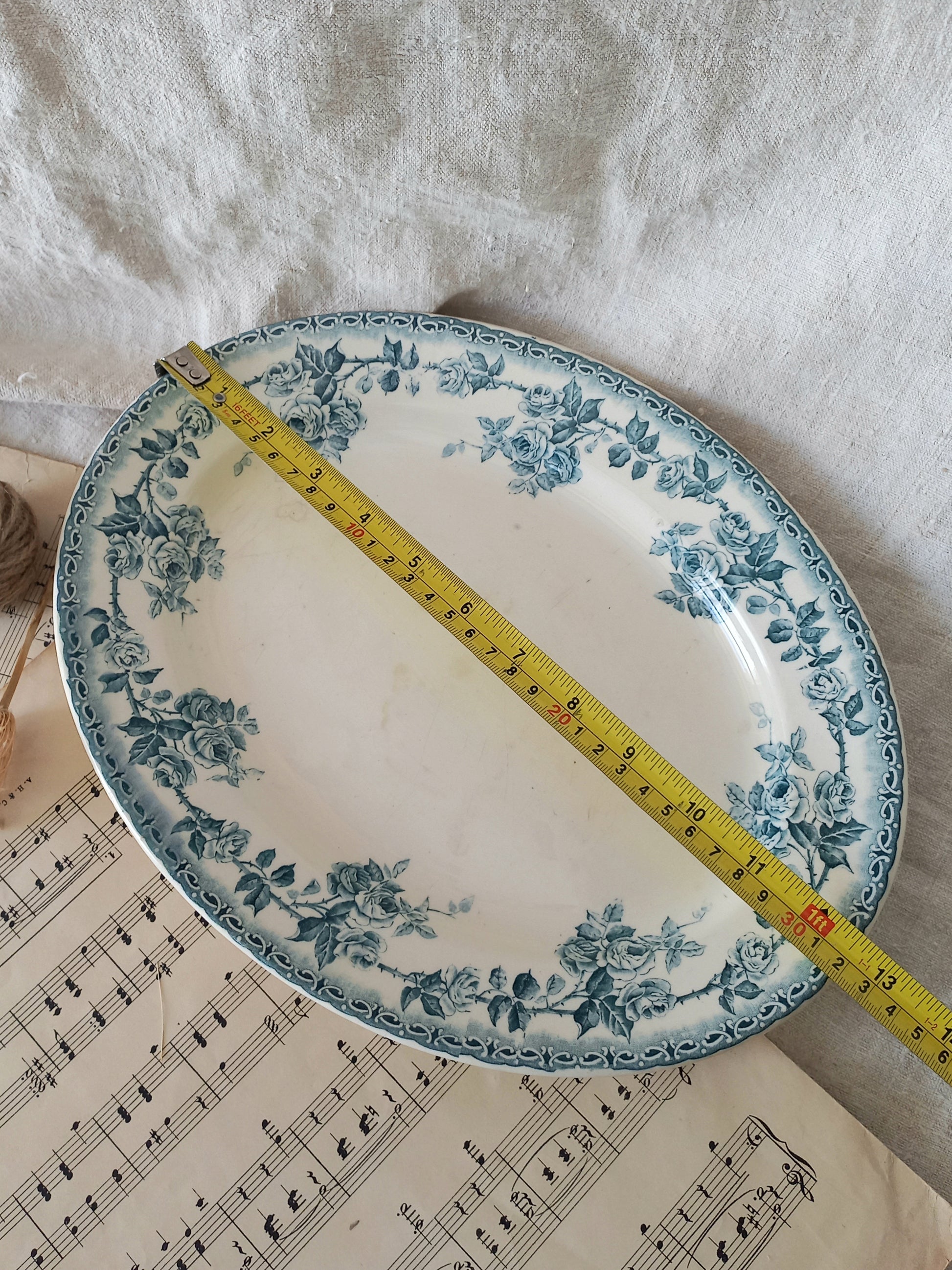 An oval ceramic serving platter with a blue floral pattern on a white background, shown with a yellow tape measure laid diagonally across it on a white fabric surface with sheet music underneath.
