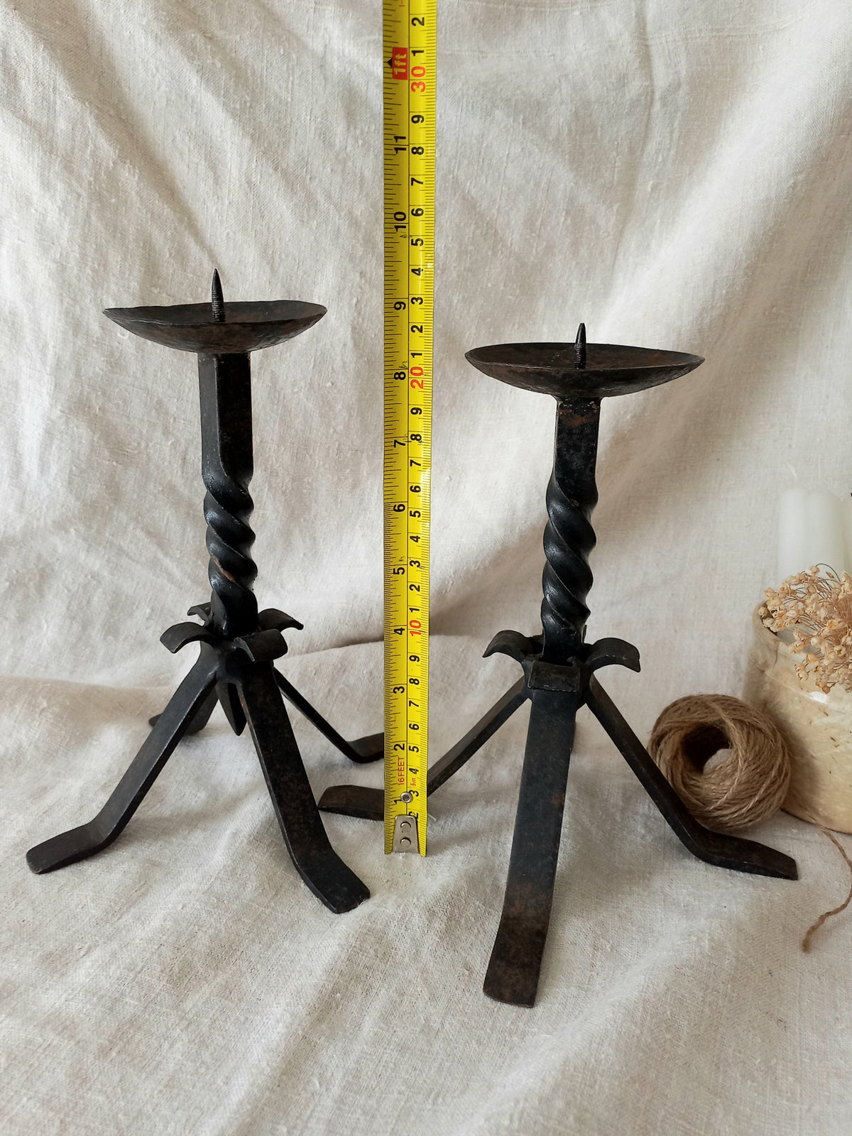 Pair of black twisted metal candle holders with a measuring tape for scale on a white fabric background.