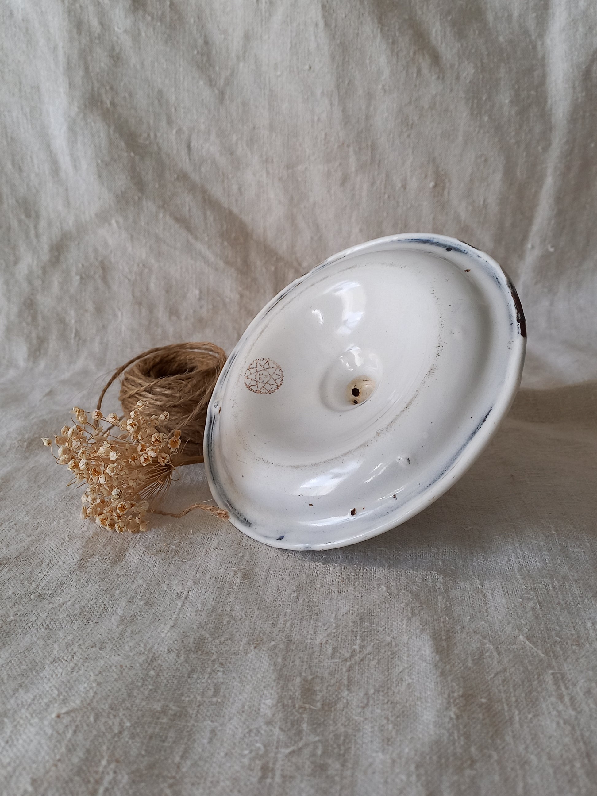 Underside of a white enamel candle holder with blue rim on a textured beige surface with twine and dried flowers.