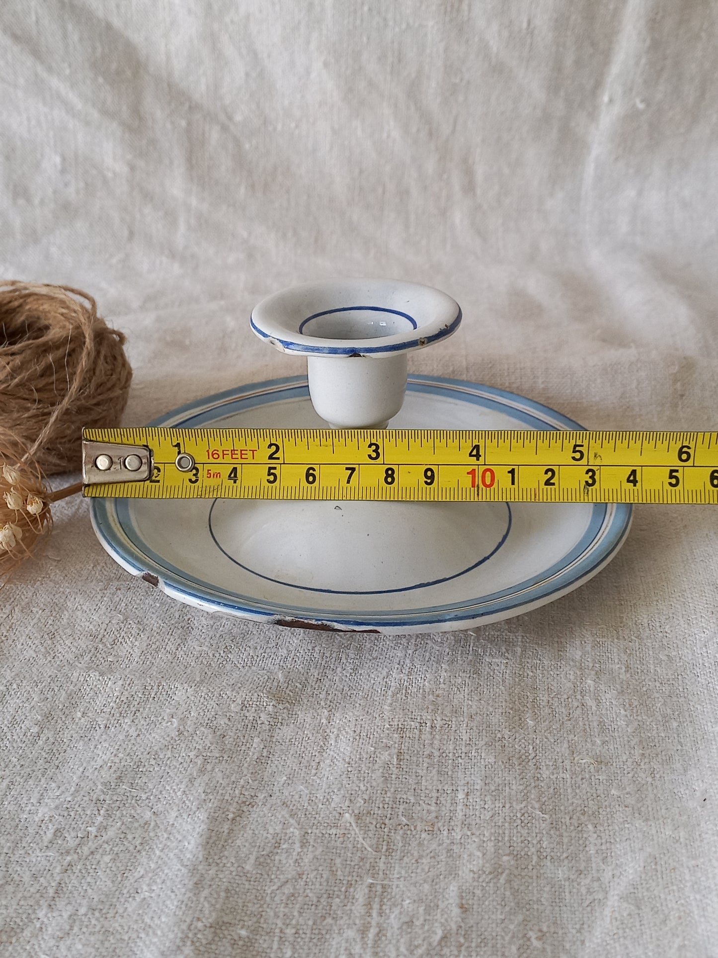 White enamel candle holder with blue rim on a textured beige surface with twine and dried flowers and a tape-measure for reference.