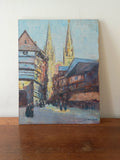 An impressionist-style oil painting of a European street scene with two tall church spires, housed buildings, and small figures, displayed on a wooden surface against a light wall.