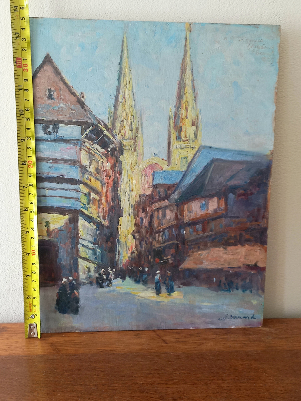 An impressionist-style oil painting of a European street scene with two cathedral spires, shown leaning against a wall on a wooden surface with a yellow tape measure along the left edge.