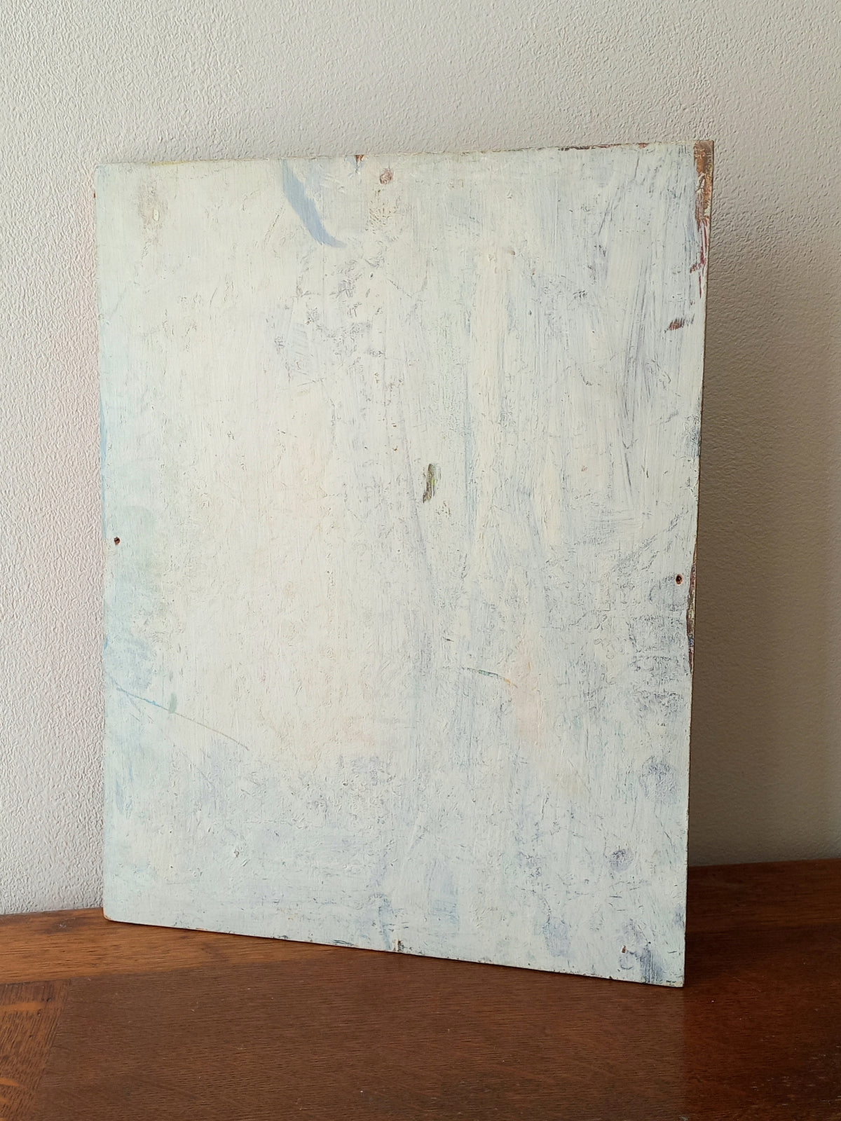 A square wooden board with a distressed, textured off-white and light blue paint finish, standing on a dark brown wooden surface against a light beige wall.