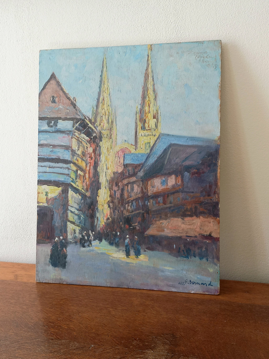 An impressionist-style painting of a European street scene with church spires, mounted on a wooden surface against a light wall.