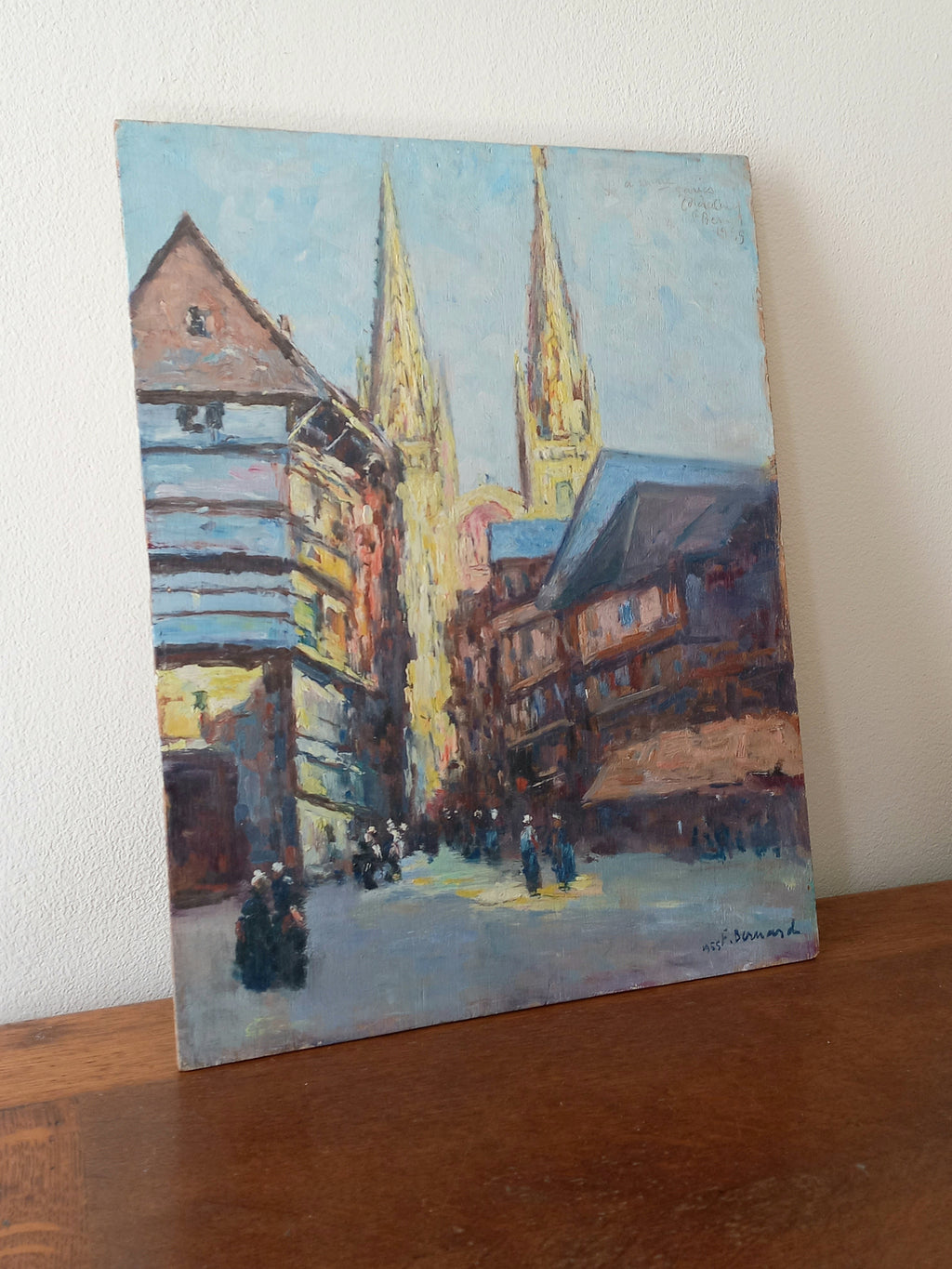 An impressionist-style painting of a European street scene with church spires, shown leaning against a white wall on a wooden surface.