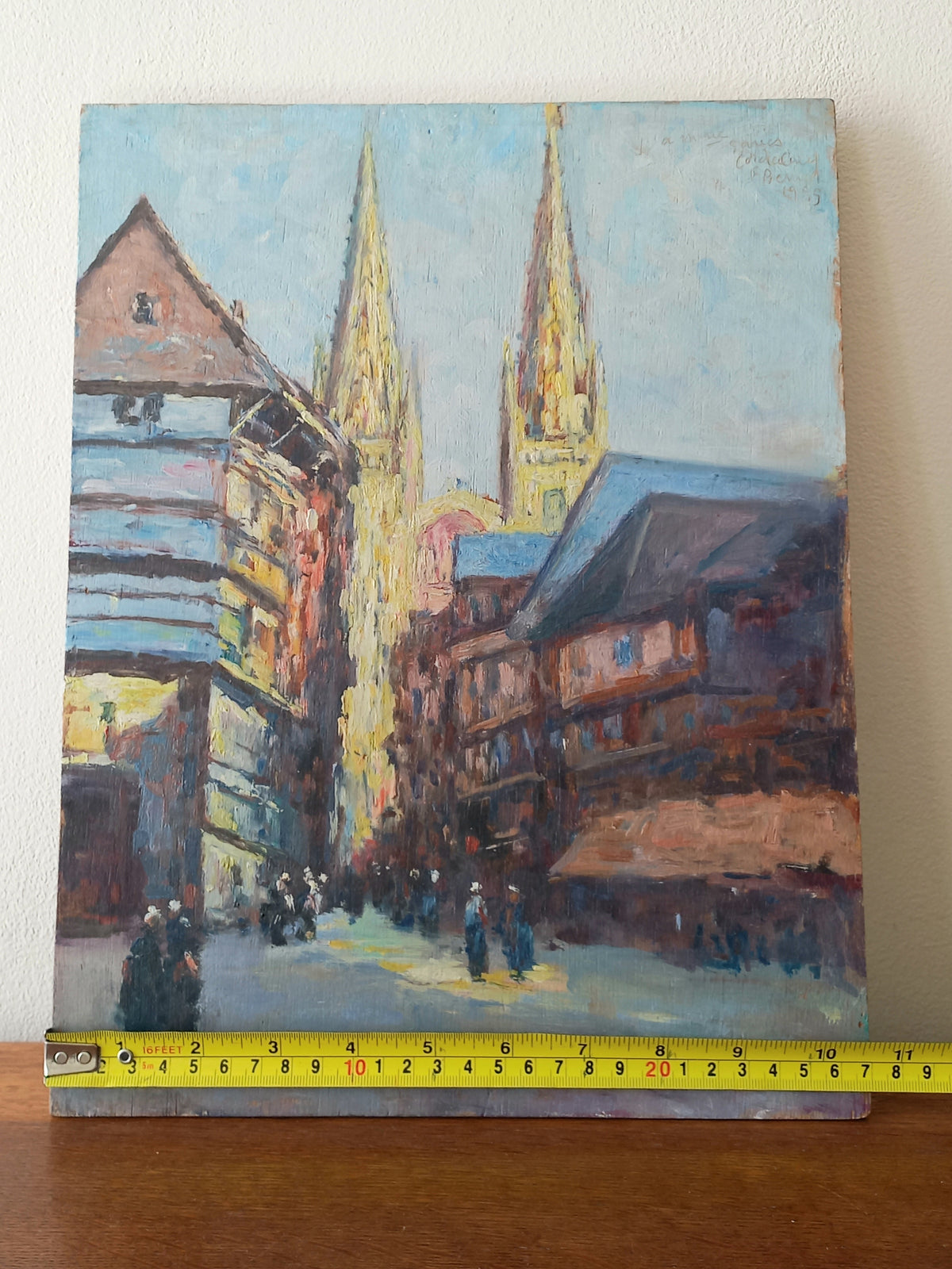 An impressionist-style painting of a street scene with two tall church spires, housed in warm-toned buildings under a pale blue sky. A yellow tape measure is placed along the bottom edge of the canvas against a light wall and wooden surface.