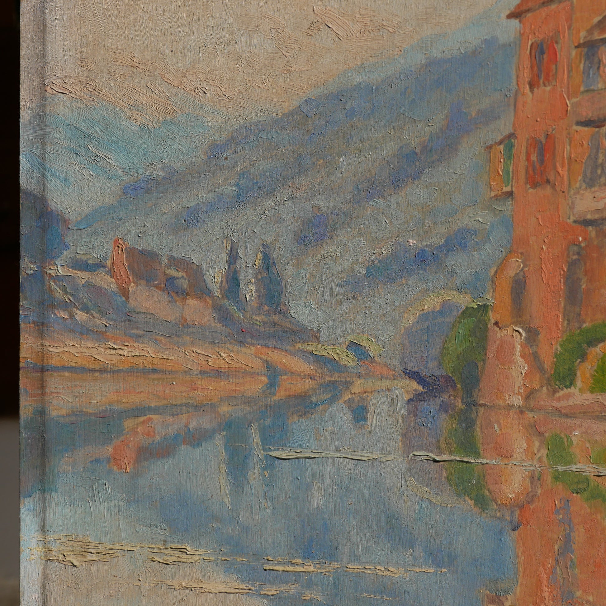 An original vintage oil painting depicting a village on the banks of a large river. It looks similar to the Rhine River Danube in Germany, and with the large hills/mountains in the background maybe the Black Forest, although it could be many places in Europe and perhaps even in France where we purchased this wonderful piece of original art on wooden board.