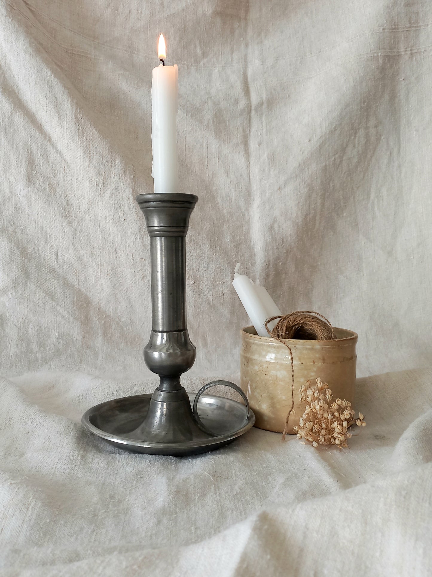 pewter candlestick holder with a lit candle on a textured white fabric background